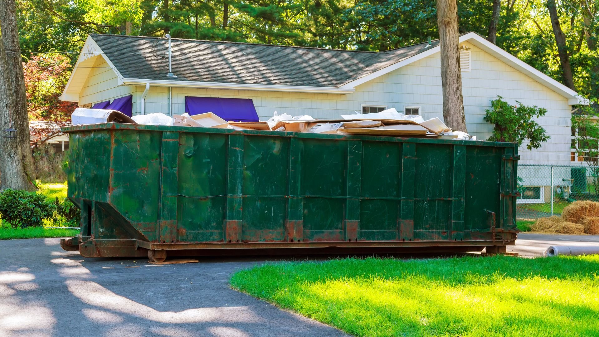 Large green dumpster filled with debris in residential driveway near white house