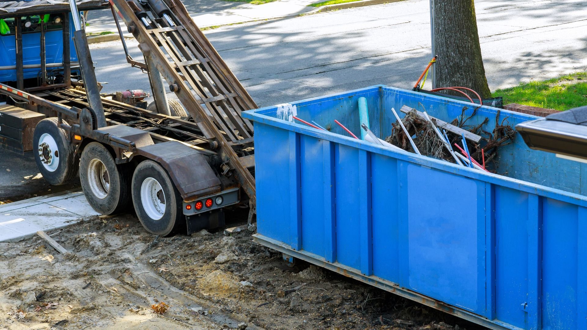 Blue dumpster and truck on dirt ground near tree during construction cleanup