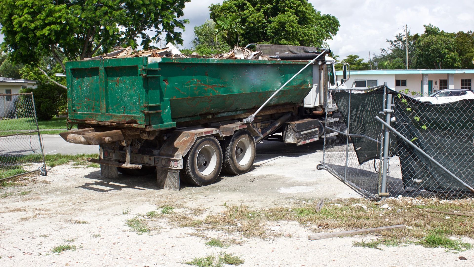 Green dumpster truck loaded with debris parked near chain-link fence