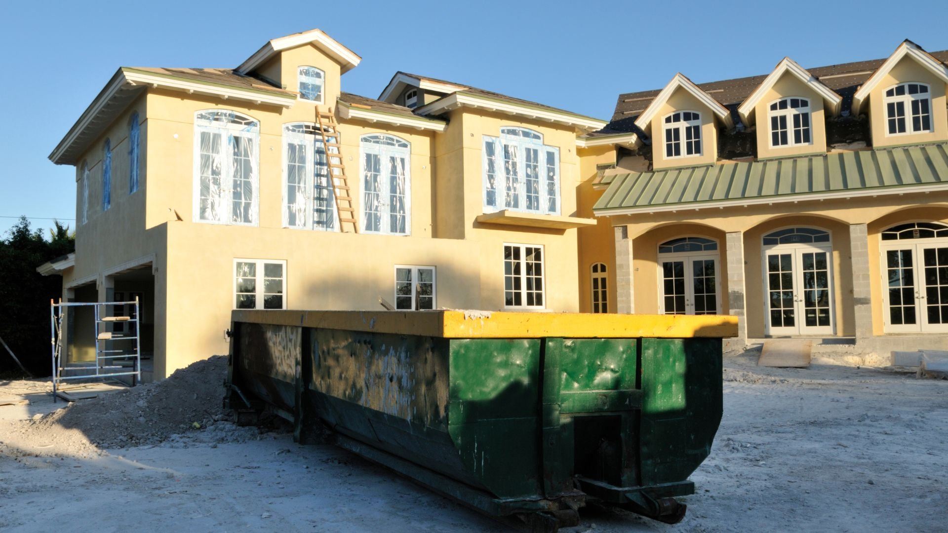Yellow house under construction with large green dumpster in foreground